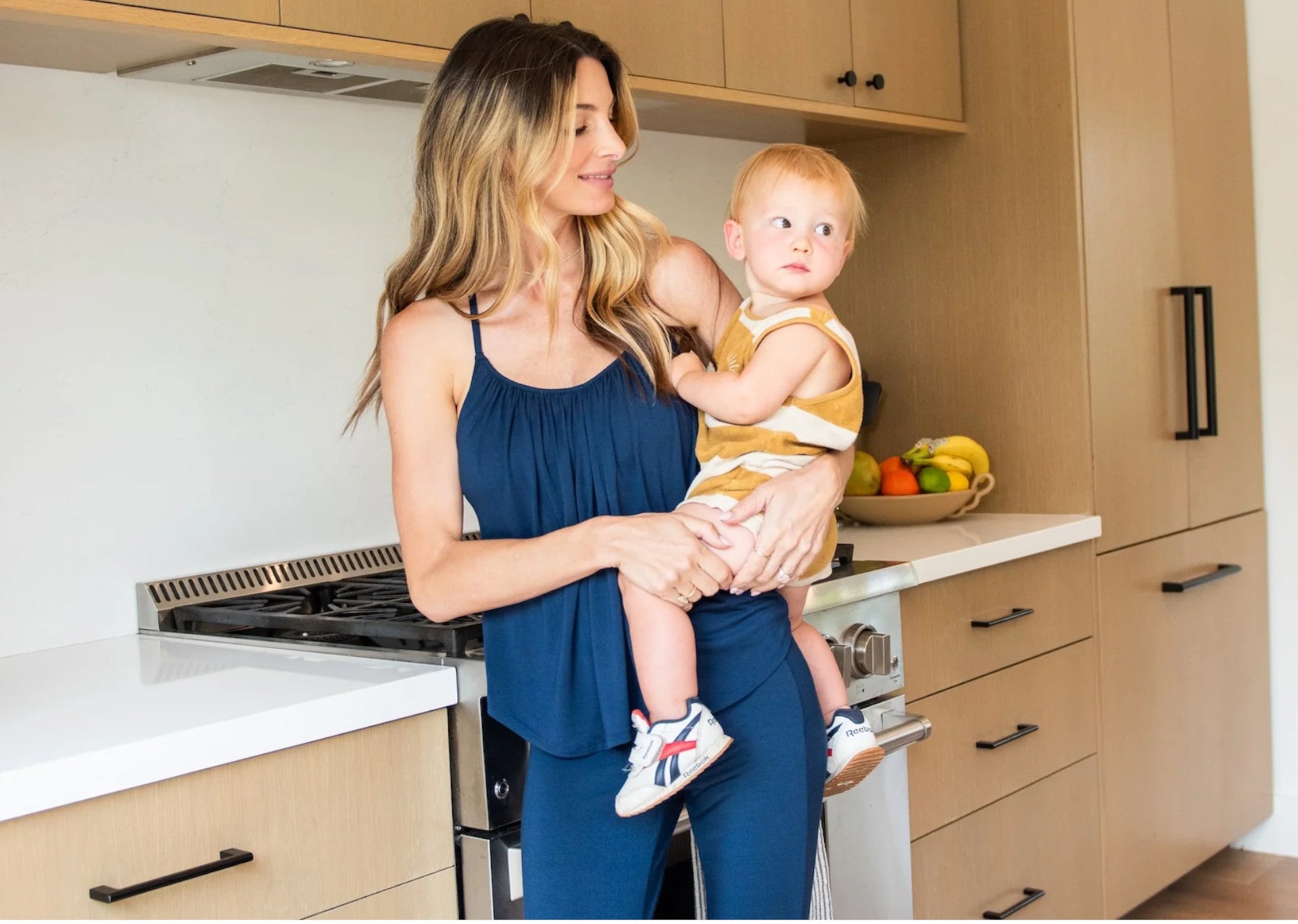 Woman in navy bra free Slumber Party top holding baby in kitchen.